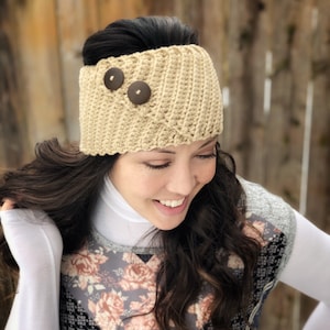 May include: A beige crocheted headband with two brown buttons. The headband is worn by a person with long brown hair.