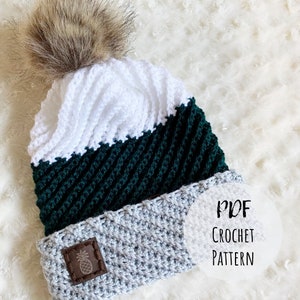 May include: Crochet hat pattern PDF download. The hat is white, green, and gray with a faux fur pom pom on top.