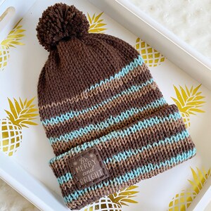 Puede incluir: Gorro de punto de rayas marrones y azules con un pompón marrón en la parte superior. El gorro tiene un parche marrón con el texto "Be Merry and Bright" en él.