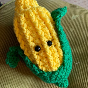 May include: A crocheted corn on the cob plush toy with green leaves and black button eyes.