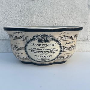 May include: A cream-colored ceramic bowl with a dark rim, featuring black musical staff lines and notes printed across the exterior. A central oval label displays the text GRAND CONCERT, LE CLAVECIN, FORTE-PIANO, and W. A. MOZART in black lettering.
