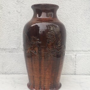 May include: A tall, brown ceramic vase with a glossy finish. The vase features embossed winged dragon designs on the front. The vase has a wide base, a cylindrical body, and a narrow neck. The vase is approximately 12 inches tall.