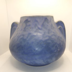 May include: A blue ceramic vase with two handles. The vase has a rounded body and a textured surface. The color is a muted blue with some lighter areas, giving it a slightly aged appearance. The handles are a lighter shade of blue.