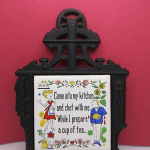 May include: Black cast iron trivet with a white ceramic tile insert. The tile features a colorful illustration of a woman and a man with the text "Come into my kitchen and chat with me While I prepare a cup of tea."
