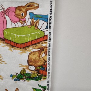May include: White fabric with a repeating pattern of brown rabbits. The rabbits are depicted in various scenes, including one rabbit tucking another rabbit into bed, and another rabbit carrying a basket of berries. The fabric has a text label that reads "Guaranteed Washable Colors Preshrunk Protected by Triple Shield - Copr. 1971,  Artcraft Products, Inc., Jersey City, N.J."