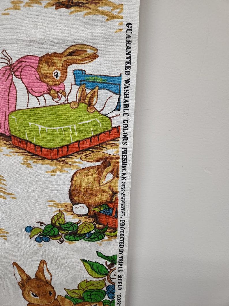 May include: White fabric with a repeating pattern of brown rabbits. The rabbits are depicted in various scenes, including one rabbit tucking another rabbit into bed, and another rabbit carrying a basket of berries. The fabric has a text label that reads "Guaranteed Washable Colors Preshrunk Protected by Triple Shield - Copr. 1971,  Artcraft Products, Inc., Jersey City, N.J."