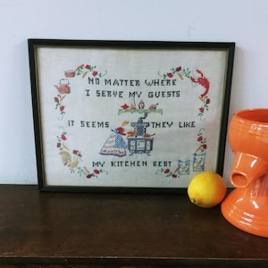 May include: A framed cross-stitch sampler with a black frame. The sampler features a woman cooking in a kitchen with the text "No matter where I serve my guests it seems they like my kitchen best".