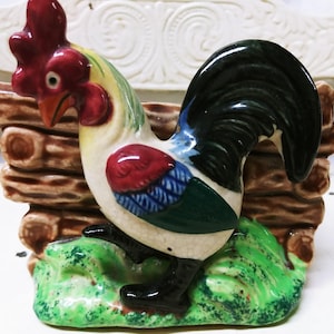 May include: A ceramic rooster figurine with a red comb, beak, and wattle, and a black tail. The rooster is perched on a green base with a brown log fence behind it. The rooster's body is white with red, blue, and green accents.