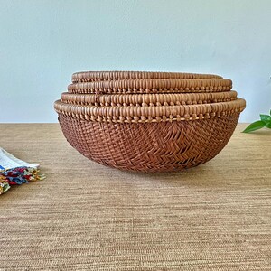 May include: A set of three woven brown baskets of varying sizes, stacked together. The baskets have a rounded shape and are made of a natural fiber. A colorful cloth with a fringed edge is on the left.