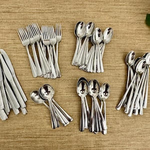 May include: A collection of stainless steel silverware, including forks, knives, and spoons, arranged on a wooden surface. The silverware is shiny and reflects light, suggesting a formal dining setting. The arrangement includes various spoon sizes.