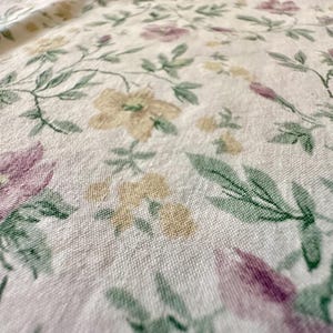 May include: Close-up of a floral fabric with a cream background. The fabric features a pattern of flowers in shades of purple, yellow, and green. The flowers are interspersed with green leaves and stems, creating a delicate and natural design.