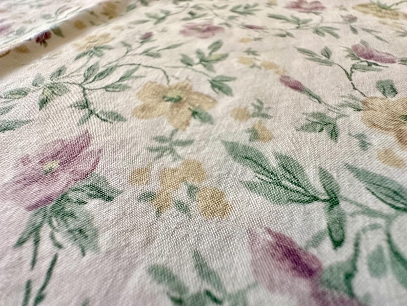 May include: Close-up of a floral fabric with a cream background. The fabric features a pattern of flowers in shades of purple, yellow, and green. The flowers are interspersed with green leaves and stems, creating a delicate and natural design.