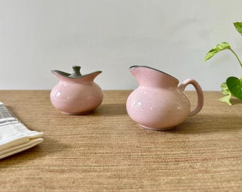 May include: A light pink ceramic creamer and a matching lidded sugar bowl, both featuring a speckled finish and a smooth, rounded shape with a grey interior rim. The creamer has a curved handle and a pouring spout, while the sugar bowl has a lid.