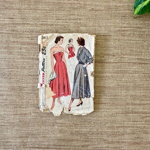 May include: Vintage sewing pattern featuring illustrations of dresses. The pattern packet is off-white with the number "2817" and the price "25c" visible. The dresses are in red and gray, with a retro style.