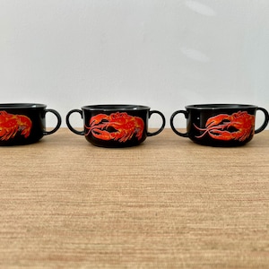 May include: Three black soup bowls with red lobster illustrations. Each bowl has two handles and a glossy finish. The bowls are arranged on a light brown surface, with a green plant in the background.
