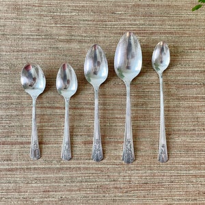 May include: Five silver spoons of varying sizes, arranged in a row on a brown woven surface. The spoons have ornate designs on the handles.