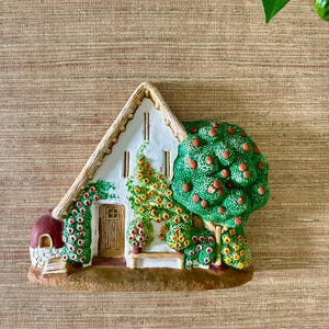 May include: A decorative ceramic cottage with a white facade, a brown door, and a red roof. The cottage is surrounded by green trees with red and yellow accents, and a small bench. The base is brown.