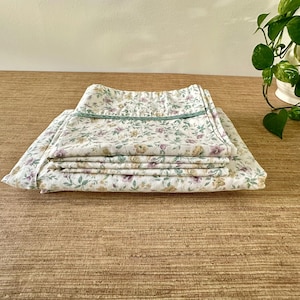May include: Folded floral bed linens with a white background and a pattern of purple, yellow, and green flowers. The top edge is trimmed with a green border. The linens are stacked on a textured, light brown surface.