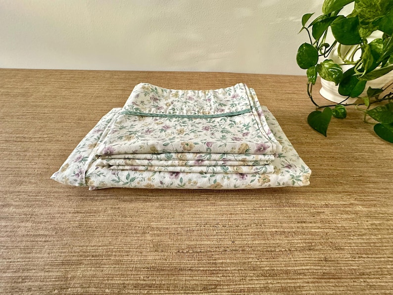May include: Folded floral bed linens with a white background and a pattern of purple, yellow, and green flowers. The top edge is trimmed with a green border. The linens are stacked on a textured, light brown surface.