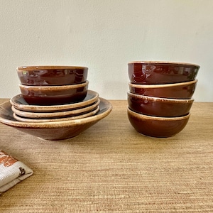 May include: A collection of brown ceramic bowls and plates. The bowls are stacked in two groups, with a stack of plates to the left. The ceramic pieces have a glossy finish and a warm, earthy tone, perfect for serving food.