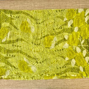 May include: A rectangular, lime green scarf with a floral pattern and a checkered border. The scarf features a repeating pattern of stylized flowers and leaves in shades of green and white. The fabric appears to be lightweight and slightly textured.