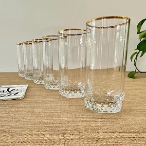 May include: A set of seven clear drinking glasses with gold rims. The glasses have a diamond-cut pattern at the base and vertical lines on the sides. A coaster with the word "drink" is in the foreground. The glasses are arranged on a brown surface.