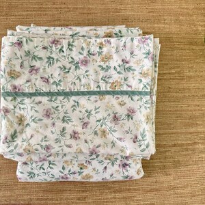 May include: Folded white floral bed sheets with a pattern of purple and yellow flowers and green leaves. A green stripe runs across the top of the sheets. The sheets are stacked on a wooden surface.