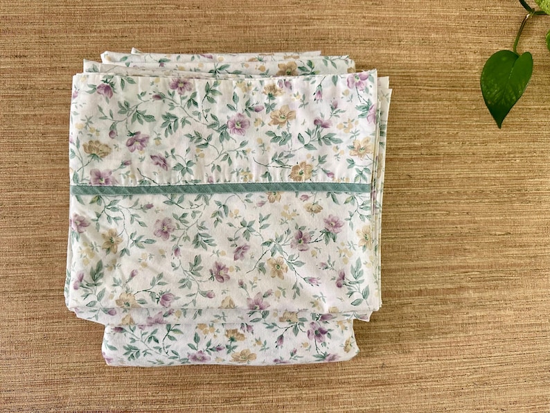 May include: Folded white floral bed sheets with a pattern of purple and yellow flowers and green leaves. A green stripe runs across the top of the sheets. The sheets are stacked on a wooden surface.