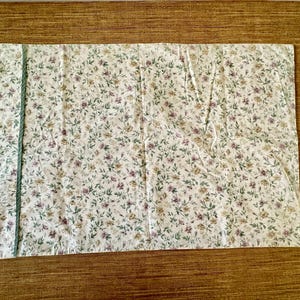 May include: A rectangular pillowcase with a floral design. The pillowcase has a white base with small purple, yellow, and green flowers and leaves. A green trim runs along one side. It is likely for a bed.