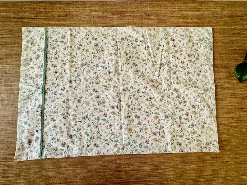 May include: A rectangular pillowcase with a floral design. The pillowcase has a white base with small purple, yellow, and green flowers and leaves. A green trim runs along one side. It is likely for a bed.