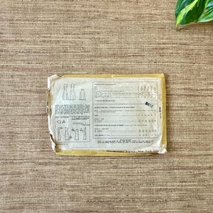May include: A vintage sewing pattern envelope with illustrations of dresses and text. The envelope is yellowed with age and has a torn edge. The background is a textured, neutral-toned surface.
