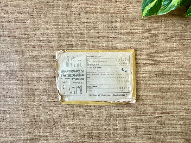 May include: A vintage sewing pattern envelope with illustrations of dresses and text. The envelope is yellowed with age and has a torn edge. The background is a textured, neutral-toned surface.