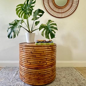 May include: A cylindrical side table constructed from stacked brown rattan reeds. A white textured pot holds a variegated monstera plant with green and white leaves. Three books are stacked on the table surface beneath the plant.