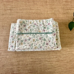 May include: Folded floral fabric with a white background and a pattern of small purple, yellow, and green flowers and leaves. A green stripe runs across the top. The fabric is on a wooden surface.