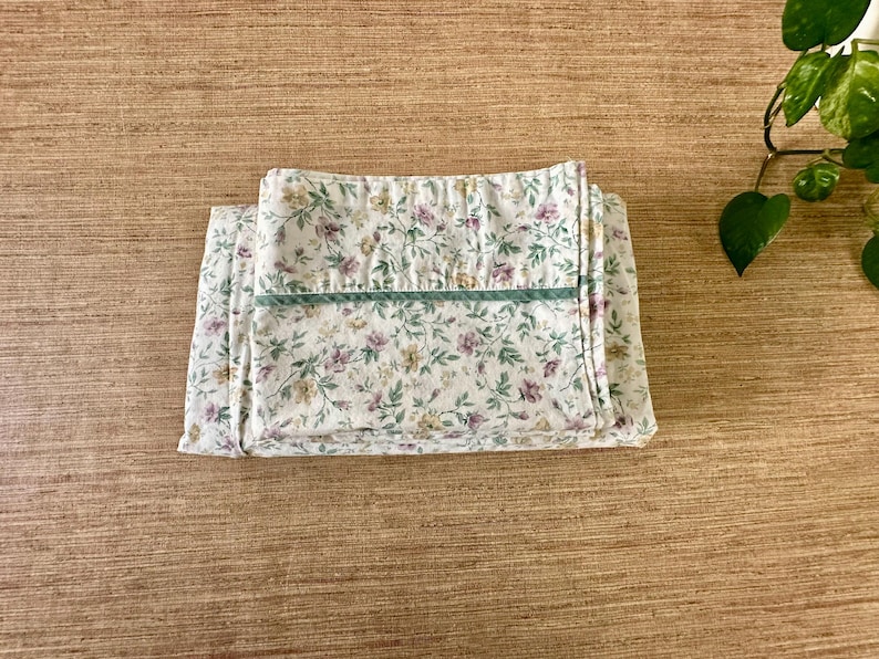 May include: Folded floral fabric with a white background and a pattern of small purple, yellow, and green flowers and leaves. A green stripe runs across the top. The fabric is on a wooden surface.