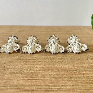 May include: Four white decorative cabinet pulls with a classical face design and dark brown accents. The pulls are arranged in a row on a light brown surface. The pulls have a distressed, vintage appearance.