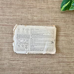 May include: A vintage sewing pattern, featuring illustrations of dresses and garment pieces. The pattern is printed on aged, off-white paper with detailed instructions and measurements. The paper is slightly torn and worn, suggesting its age.