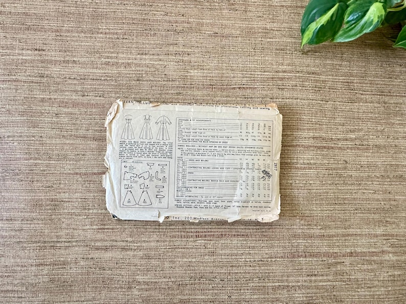 May include: A vintage sewing pattern, featuring illustrations of dresses and garment pieces. The pattern is printed on aged, off-white paper with detailed instructions and measurements. The paper is slightly torn and worn, suggesting its age.