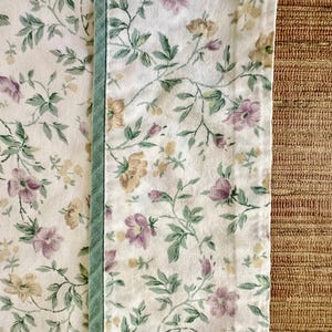 May include: Floral fabric with a pattern of purple, yellow, and green flowers and leaves on a white background. A vertical strip of light green fabric is sewn along the edge. The fabric is likely for home decor.