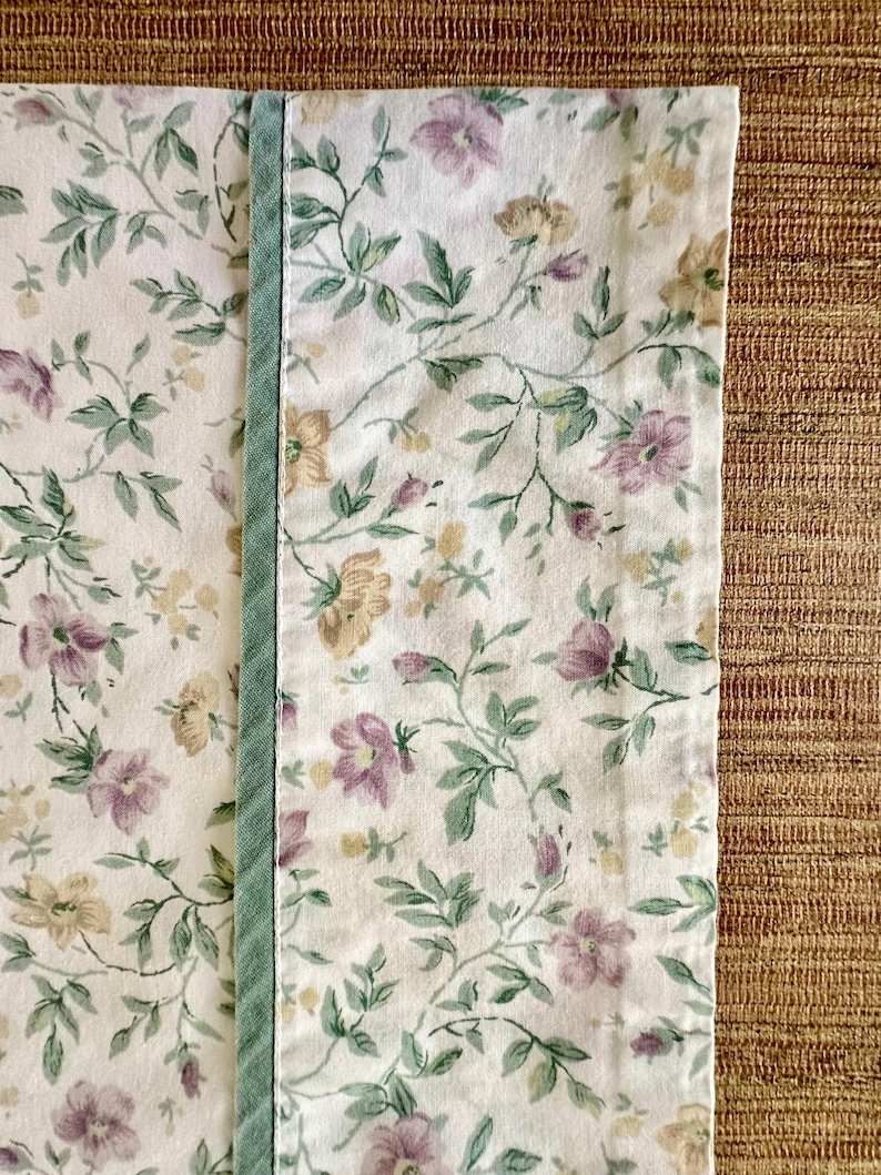 May include: Floral fabric with a pattern of purple, yellow, and green flowers and leaves on a white background. A vertical strip of light green fabric is sewn along the edge. The fabric is likely for home decor.
