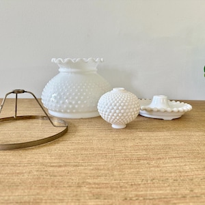 May include: A collection of white glass objects, including a hobnail-textured lamp shade, a small hobnail-textured vase, a candle holder, and a metal lamp frame. The objects are arranged on a light brown wooden surface.