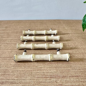 May include: Four bamboo-style drawer pulls with a distressed, off-white finish. Each pull has a cylindrical shape with dark accents, resembling bamboo stalks. The pulls are arranged on a textured, brown surface.