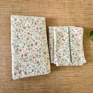 May include: A floral bedding set, comprising a pillowcase and two sheets, folded neatly. The fabric has a white base with a repeating pattern of small purple, yellow, and green flowers and leaves. One sheet is accented with a green stripe.