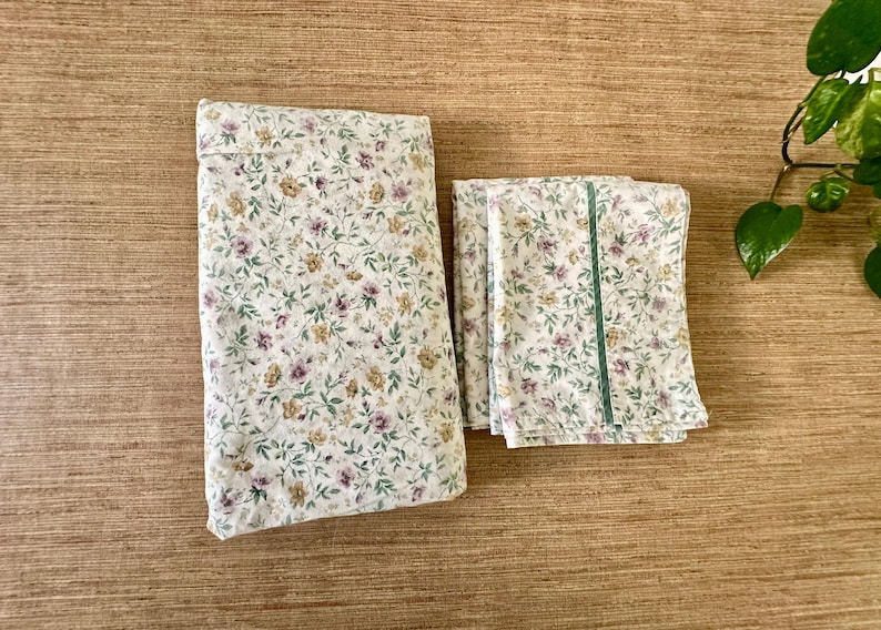 May include: A floral bedding set, comprising a pillowcase and two sheets, folded neatly. The fabric has a white base with a repeating pattern of small purple, yellow, and green flowers and leaves. One sheet is accented with a green stripe.