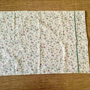 May include: A rectangular piece of fabric with a floral pattern on a cream background. The pattern includes small purple, yellow, and green flowers and leaves. A thin, green stripe runs along one edge. Likely a pillowcase or similar textile.