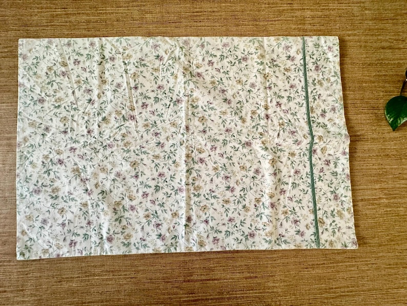 May include: A rectangular piece of fabric with a floral pattern on a cream background. The pattern includes small purple, yellow, and green flowers and leaves. A thin, green stripe runs along one edge. Likely a pillowcase or similar textile.