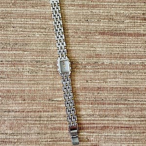 May include: Silver-toned wristwatch with a rectangular face and a linked band. The watch has a light-colored face with small markers. The band is made of silver links with gold-colored accents. The watch is displayed on a textured, brown surface.