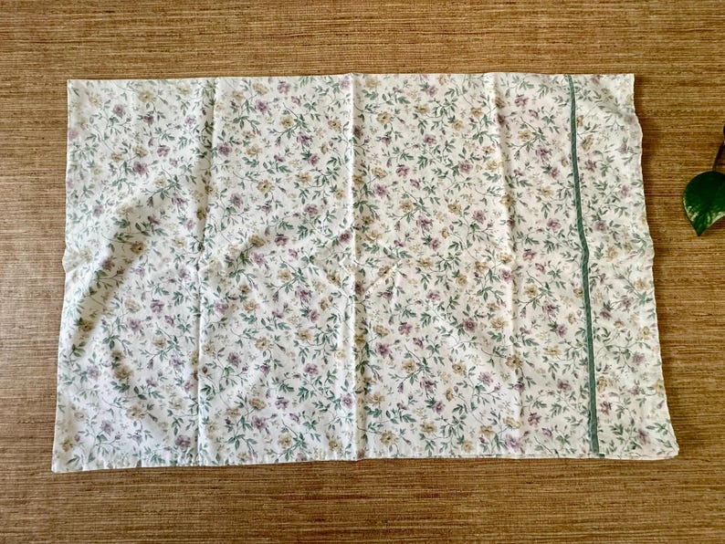 May include: White pillowcase with a floral pattern of small purple, yellow, and green flowers and leaves. A thin green stripe runs vertically along one side. The pillowcase is made of a soft, lightweight fabric.