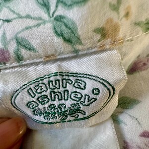 May include: Close-up of a white fabric with a floral pattern in green, purple, and yellow. A white label with the green text "Laura Ashley" is visible. The fabric is likely part of a garment or textile.