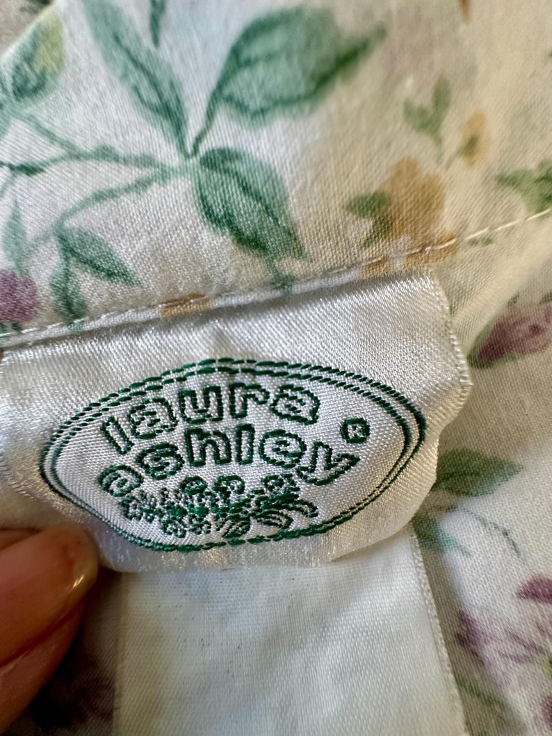 May include: Close-up of a white fabric with a floral pattern in green, purple, and yellow. A white label with the green text "Laura Ashley" is visible. The fabric is likely part of a garment or textile.